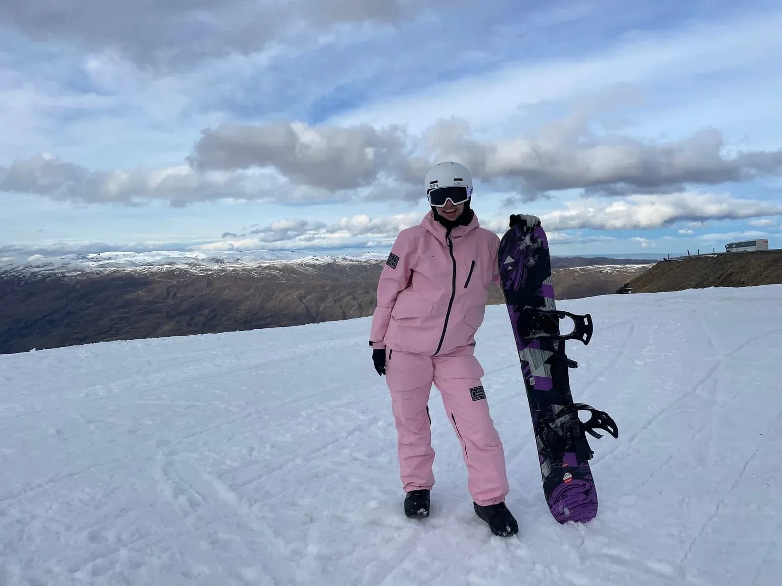 A snowboarder in pink stands with a purple board against a snowy mountain landscape.