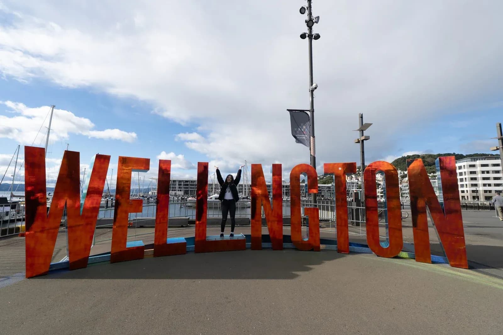 A visitor stands among large orange Wellington letters beside boats on the waterfront.